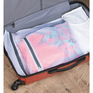 An open red suitcase lies on a wooden floor. Inside, clothes are neatly packed, including a floral garment in an M Design Laundry & Travel Storage Zipper Bag (3-Pack) for delicate protection and a white lacy item. The suitcase features a grey interior.