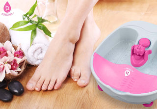 A pair of bare feet rests on a wooden floor beside the Pursonic Foot Spa Massager with Tea Tree Oil Soak, designed for ultimate relaxation. Nearby, spa accessories including rolled towels, black stones, and pink flowers in a coconut shell create a soothing atmosphere. The background features bamboo leaves, tea tree oil bottles from Pursonic, and another white rolled towel.