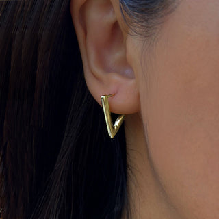 Close-up of a woman's ear wearing the Achieve Triangle Huggie Earrings by Sonia Hou Jewelry. These modern, minimalist gold earrings feature a V-shaped design reminiscent of triangle earrings. The sleek and polished finish serves as a motivational reminder to embrace change, contrasting beautifully with the darker tones of her hair and skin.