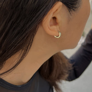 Close-up view of a person with dark hair wearing a small, crescent-shaped gold earring from Sonia Hou Jewelry's Essential Huggies collection. The lightweight, versatile earring is visible on their left ear as their head is turned to the right. The background is blurred.