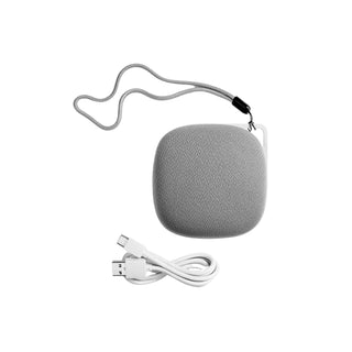 The Yogasleep Travelcube Portable Sound Machine, a grey Bluetooth speaker with a lanyard attached, sits against a white background with a white USB charging cable placed below it. The speaker features a fabric texture and compact, rounded square shape—making it an ideal travel essential for on-the-go relaxation.