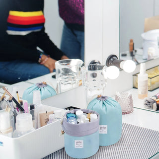 A vanity table with various makeup and skincare products neatly organized. Two light-blue Waterproof Makeup Organizer Travel Bags (2-Pack) from Amore Paris hold items alongside a white basket filled with brushes and bottles. A large mirror with lit bulbs reflects a person wearing a colorful striped top.