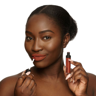 A person with smooth, dark skin applies a matte red lipstick from the Mented Blood Orange Liquid Lipstick (2-Pack) while holding the container in their other hand. They have neatly styled hair pulled back. The makeup accentuates their glowing complexion and they are smiling slightly.
