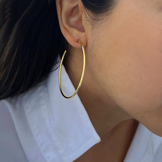 A close-up of a person wearing Sonia Hou Jewelry's Perfect Hoop Earrings. The person has long, dark hair and is dressed in a white collared shirt. The focus is on the large, gold hoop earring and part of the ear, with the rest of the face not fully visible.