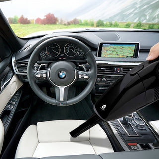 A person uses the Pursonic Handheld Car Vacuum Cleaner (with Adapter Included) featuring 1800PA super suction to clean the interior of a car. The car's dashboard and GPS system are visible, along with the steering wheel and gear shift. The interior is black and white, and there's a scenic landscape visible through the windows.