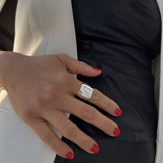 A close-up of a person's hand resting on their hip. The individual is styled in a sophisticated black outfit paired with a white blazer—a true wardrobe essential. Their red nails are impeccably manicured, and they are making a grand statement with Sonia Hou Jewelry's Rich Chunky Wide Cigar Band Ring adorning their middle finger, its shiny silver surface gleaming prominently.