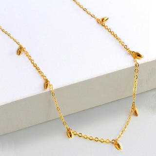 A delicate gold necklace featuring small, rice bead-shaped pendants hanging at intervals along a fine chain. The Minimalist Rice Bead Chain Necklace by Sonia Hou Jewelry symbolizes wealth and prosperity, displayed on a white surface to highlight its elegant and simple design.