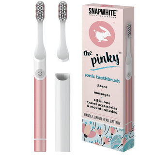 A pink and white electric toothbrush beside its packaging. The box displays the brand name "Snapwhite" and product name "The Pinky" Electric Travel Toothbrush, accompanied by two additional brush heads. The text highlights features such as superior cleaning performance, massaging, travel accessories, and an included mount.