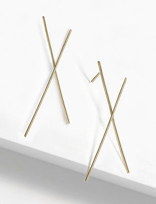 Minimalistic Chopstick Earrings by Sonia Hou Jewelry, composed of two thin, crisscrossed gold rods on a white surface. Designed to resemble abstract crosses or X shapes, they create a clean and modern aesthetic that subtly celebrates the spirit of teamwork and cooperation.